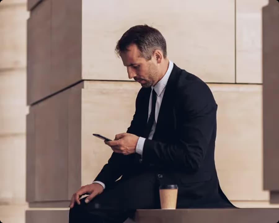 Geschäftsmann in schwarzem Anzug schaut auf sein Smartphone mit Kaffee